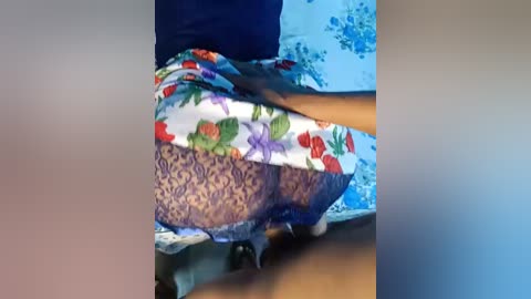 A close-up video of a woman's lower body in a blue dress with floral patterns. Her legs are spread, revealing lacy purple panties. The background is blurred.