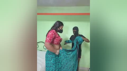 Video of two women in a room; one with long black hair, red crop top, and teal skirt, the other masked and wearing a black burqa, adjusting her teal sari.