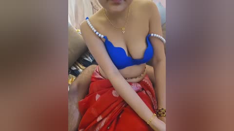Video of a light-skinned woman in a blue bra and red sari with gold jewelry, sitting indoors on a couch with a blurred background.
