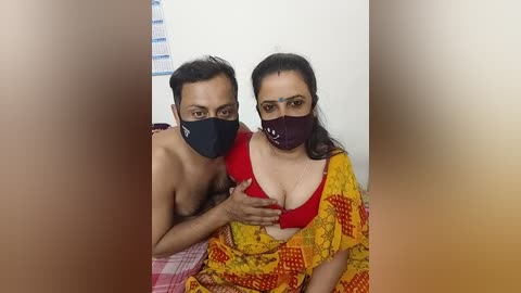 Video of a shirtless man and woman in a bedroom, both wearing masks; she has a red bra visible under a yellow sari, and he has a black mask.