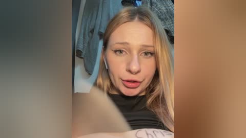 A candid video of a young woman with fair skin, blonde hair, and a black t-shirt, taken from a low angle. Background includes a messy room with clothes hanging.