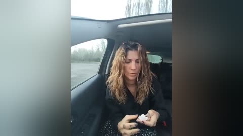 Video of a woman with wet, wavy, blonde hair, wearing a black jacket, sitting in a car, holding a white object. The background shows a foggy, overcast day.