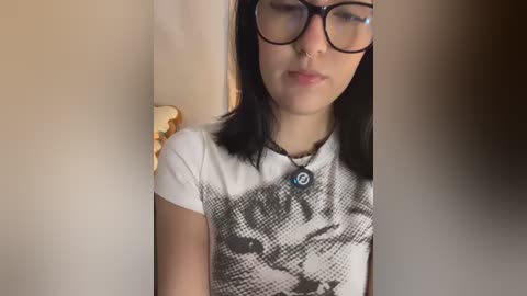 Media: Video of a young woman with straight black hair, wearing black-framed glasses and a white t-shirt with a black-and-white cat design, standing in a dimly lit room with beige walls.