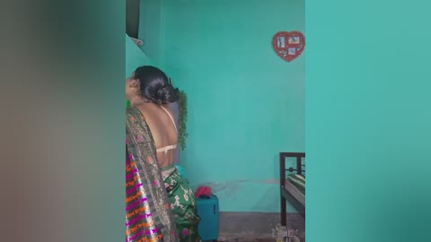 A video of a woman in a colorful sari, back to the camera, in a teal-painted room with a heart-shaped sign, a bed, and a blue container.