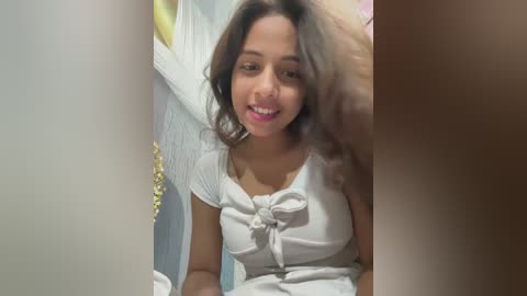 Video of a young woman with medium brown skin, long brown hair, and a white, tie-front top, smiling while being petted by a man's hand in a bathroom with a shower curtain and a towel rack in the background.