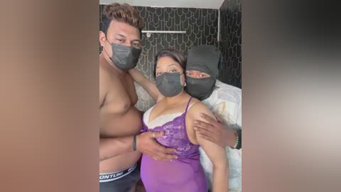Video of three masked individuals: two men and one woman, wearing purple lingerie. Background shows a bed with patterned wallpaper, and a man in white shirt.
