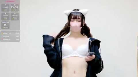 Video of an Asian woman in a white bra, black jacket, and cat ears, holding a smartphone, indoors, with a digital clock in the corner.