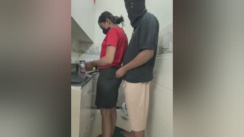 Video of a man in a black mask, dark shirt, and beige shorts holding a woman's black skirt in a small, tiled bathroom.