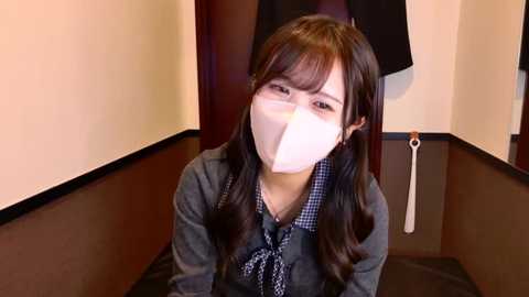 Media: Video of an Asian woman with long dark hair, wearing a face mask, blue plaid shirt, and dark jacket, sitting in a dimly lit room with beige walls and a black-and-white abstract painting.