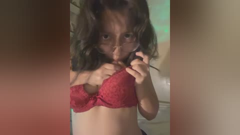 Media: Video of a young woman with medium-length dark hair, wearing a red lace bra, making a face in front of a beige wall, with blurred background.