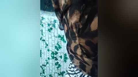 Video of a person wearing a leopard print jacket, standing in a room with a green ivy-patterned wallpaper and a white brick wall. The image is slightly blurred.