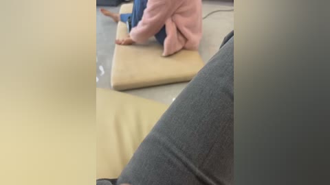 Video of a person in a pink sweater and blue jeans sitting cross-legged on a beige cushion in a room with a gray carpet. The focus is on their lower body and legs.