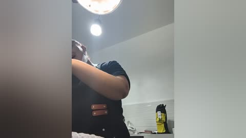 Video of a woman with medium skin tone, wearing a black shirt, standing in a dimly lit attic. She covers her face with her right arm. In the background, there is a yellow canister and a white ladder.