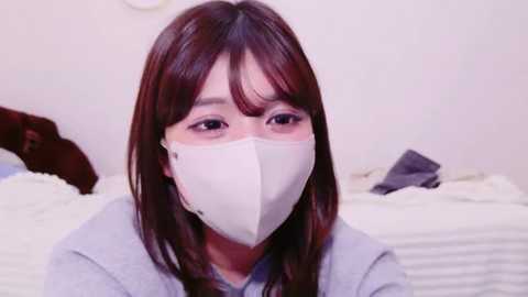 Video of an East Asian woman with long brown hair and light skin, wearing a white face mask, gray shirt, and sitting on a white bed with a dark brown headboard.