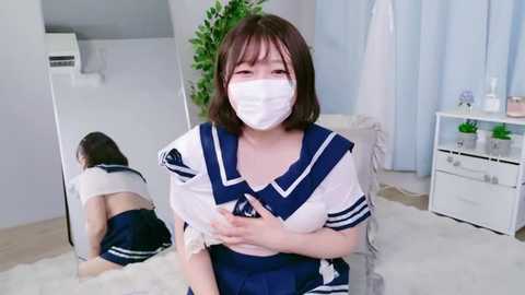 Video of an Asian woman in a schoolgirl cosplay outfit, with a white mask, kneeling on a white carpet, her reflection in a mirror showing her bent over. Background features light-colored walls and a plant.