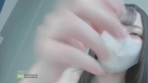 Media: A blurred video of a young Asian woman with long black hair, wearing a white face mask, and appearing to be covering her mouth with her hands.