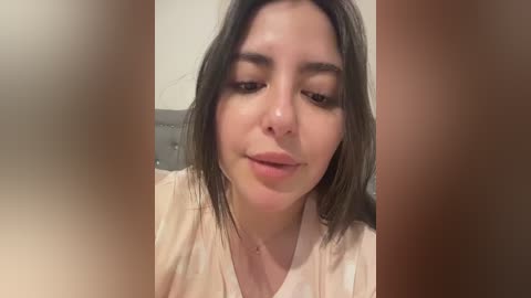 Media: A close-up video of a young woman with fair skin, straight brown hair, and brown eyes, wearing a light-colored shirt. The background is blurry, showing a grey headboard.