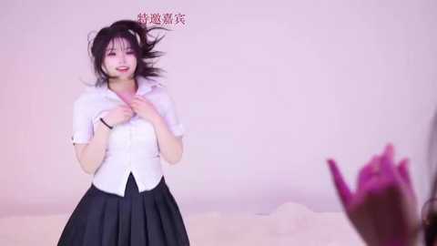 Video of a smiling East Asian woman with black hair, wearing a white blouse and black pleated skirt, standing against a pink background.