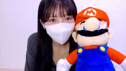 Video of an Asian woman with long black hair, wearing a white mask and dark top, hugging a plush Mario toy with an orange hat and blue overalls.