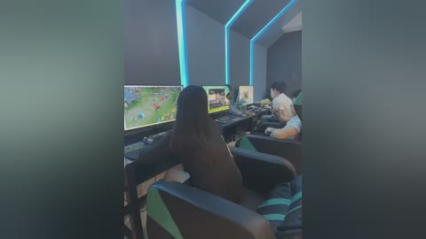 Video of a modern gaming room with two gamers, one Asian woman and one Asian man, seated in black-and-green gaming chairs. They play on multiple screens, with a vibrant game displayed. Blue LED lights accentuate the setup.