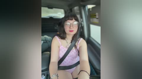 Media: A video of a young woman with pale skin, wearing glasses and a pink lace bodysuit, seated in a car with a black seatbelt across her chest.