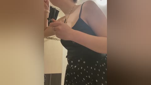 Video of a light-skinned woman in a black spaghetti-strap top, holding a phone near a beige tiled wall, with a black and white polka-dot skirt.