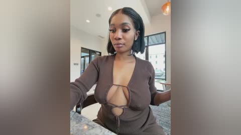 Video of a confident Black woman with a medium complexion and straight black hair, wearing a low-cut brown ribbed top, standing in a modern cafe with large windows and soft lighting.