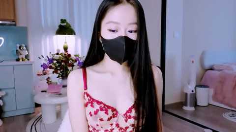Video of an East Asian woman with long black hair, wearing a red floral dress, black face mask, and a black mask, indoors, with a white table, green lamp, and cat toys in the background.