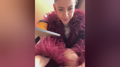 Media: Video of a young woman with medium skin tone, wearing a fluffy maroon coat, black top, and white necklace, sitting on a white table, holding a yellow cup, with blurred background.