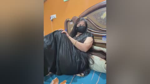 A video of a woman in a black face mask, black dress, and gloves, lying on a bed in a brightly lit room with orange walls.