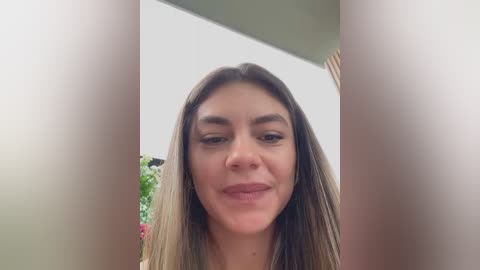 Video of a smiling woman with straight, shoulder-length brown hair, framed by two blurred vertical lines. Background includes a white wall and green plants.