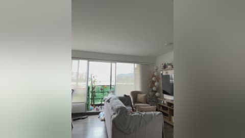 Video of a modern, minimalist living room with white walls, large floor-to-ceiling windows, a beige couch, and a flat-screen TV mounted on a wooden entertainment center. A green plant stand is visible in the background.