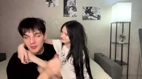 Video of a man with short black hair, wearing a black shirt, being hugged from behind by a woman with long black hair, wearing a white shirt. They're in a modern bedroom with minimalist decor.