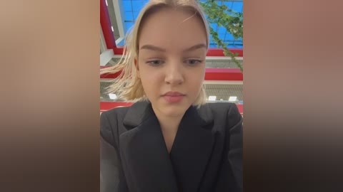 Video of a young woman with fair skin, straight blonde hair, and a neutral expression, wearing a black blazer. Background features a modern, colorful building with red and blue accents.