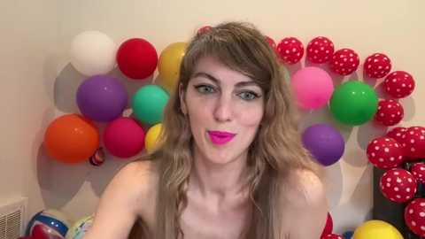 Video of a fair-skinned woman with long, wavy blonde hair, wearing vibrant pink lipstick, in a festive room adorned with colorful balloons and red polka-dotted streamers.