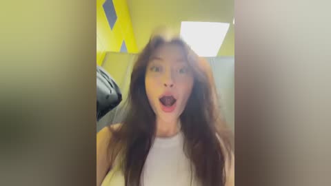 Video of a young woman with long, dark hair and light skin, wearing a white top, standing in a brightly lit room with yellow walls and a white ceiling. She has a surprised expression with her mouth open.