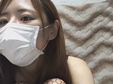 A close-up video of an East Asian woman with long, straight brown hair, wearing a white medical mask, and a beige quilted blanket in the background.