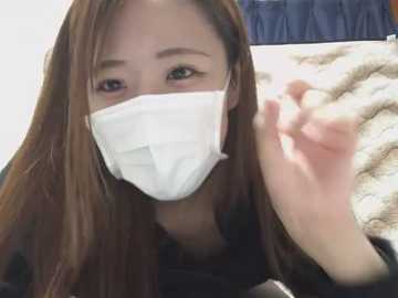 Video of an Asian woman with long brown hair, wearing a white medical mask, black top, and beige blanket, making a hand gesture, indoors.