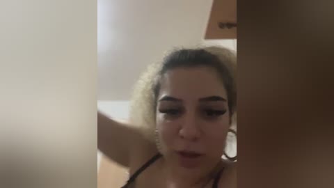 Video of a young woman with curly, blonde hair, wearing black lingerie, looking down with a neutral expression. The background is blurred, showing a wooden surface and a beige wall.