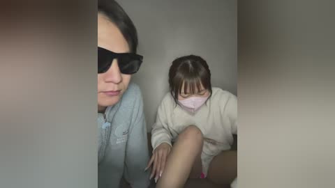 Media: Video of an Asian woman with fair skin, wearing a light blue shirt and black sunglasses, sitting beside a young Asian girl with short brown hair, wearing a light beige sweater and a pink face mask, both looking down.