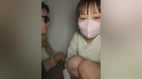 Media: Video of an Asian child with straight brown hair and pale skin wearing a pink face mask, sitting on a bed with a mirror reflecting a person wearing sunglasses.