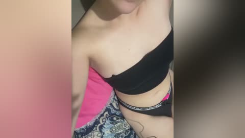 Media: A video of a light-skinned woman with a tattoo on her right hip, wearing a black bandeau and black panties with a pink waistband, lying on a bed with a blue and white patterned blanket.