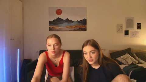 Video of two young women with light skin and long brown hair, one in a red tank top, the other in a black shirt, sitting on a dark sofa in a cozy living room with a mountain sunset painting on the wall.