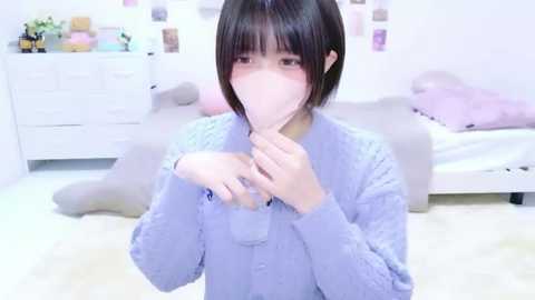 Video of an Asian woman with short black hair, wearing a light blue sweater, adjusting a pink face mask in a brightly lit, minimalist bedroom with pastel decor and white furniture.