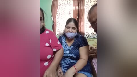 Video of a woman with medium skin tone, dark hair, and a purple face mask, wearing a blue patterned dress, seated on a couch, surrounded by others in a room with green and floral-patterned curtains.