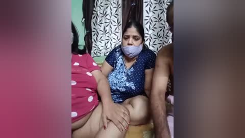 Video of an Indian woman, gagged and bound, sitting on a floor, wearing a blue patterned shirt and pink polka-dot shorts. A man, shirtless, stands behind her.