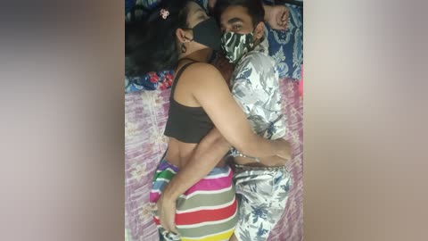 Video of two people, one in a black crop top and colorful striped shorts, embracing on a pink, floral-patterned bedspread; the other in silver foil. Both wear black masks, suggesting a protective or intimate moment.