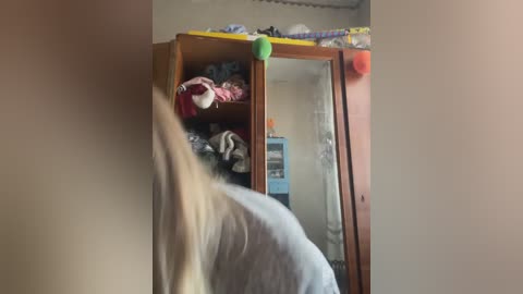 A video of a person with long blonde hair, wearing a light gray shirt, leaning over a wooden wardrobe with a mirror, displaying various colorful clothes and a blue refrigerator in the background.