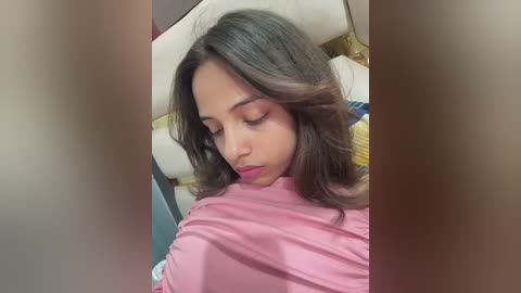 Video of a young woman with medium brown skin and long, wavy black hair, wearing a pink robe, lying on a white couch with a yellow blanket.