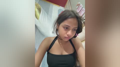 Video of a young woman with medium-brown skin and long, wavy black hair, wearing a black halter top, standing in a room with a white bed, pink and beige curtains, and gold accents.